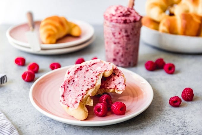 An image of a roll slathered in raspberry butter on a pink plate with fresh raspberries on the side.