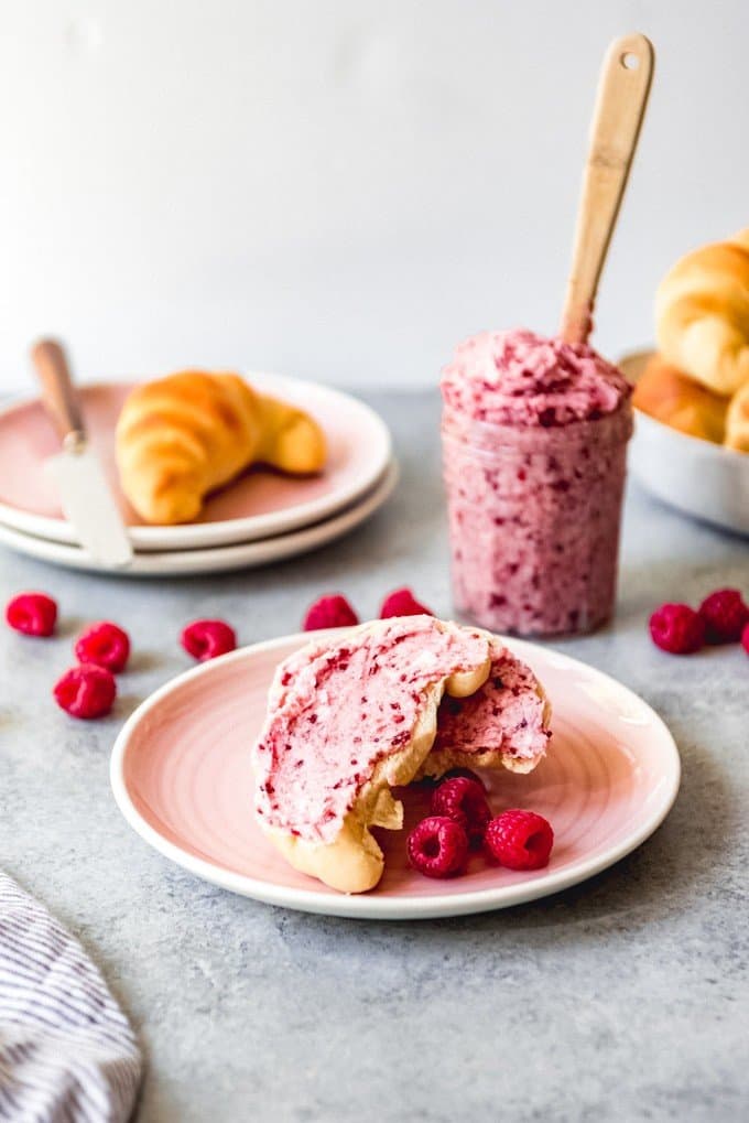 crescent rolls in a bowl and on plates with scattered fresh raspberries around and a jar of raspberry butter with a wooden handlee behind a crescent roll with butter spread on it
