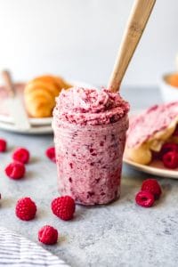 a glass jar with raspberry butter and a wooden handle inside and surrounded by scattered berries