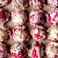 An image of balls of chocolate chip cookie dough with raspberries lined up in rows on a baking sheet.