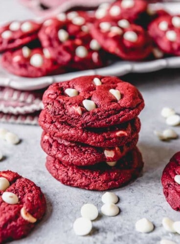 red velvet cookies with white chocolate chips stacked up with a plate of cookies in back and a cookie to each side with scattered chocolate chips everywhere