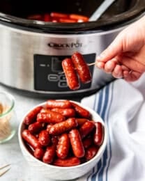 A hand holding a toothpick with two honey garlic little smokies on it.