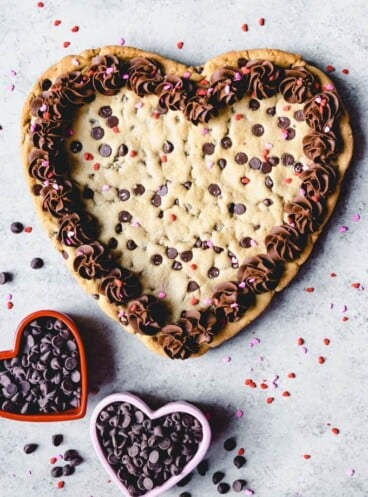An aerial view of a heart shaped giant chocolate cookie with frosting and sprinkles and heart shaped bowls filled with chocolate chips to the side