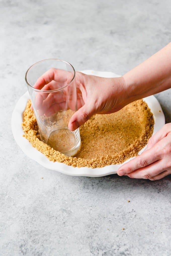 An image of a hand holding a glass to press buttery graham cracker crumbs into a graham cracker crust in a pie plate.