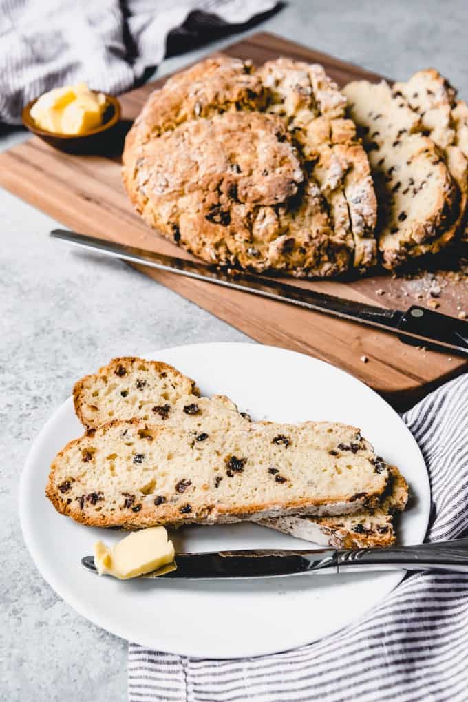 An image of two sliced of authentic Irish soda bread with currants on a white plate with Irish butter and a loaf of bread in the background.