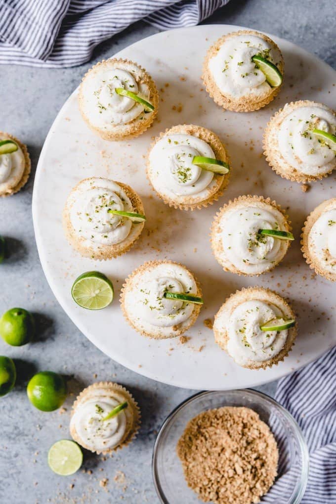 An image of key lime cupcakes with swirls of key lime buttercream frosting on a cake stand with a bowl of graham cracker crumbs and key limes next to them.