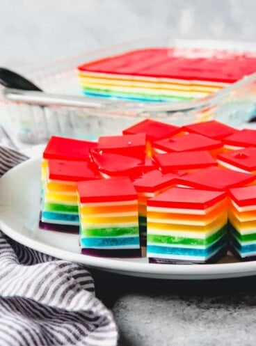 layered rainbow jello cubes on a white plate in front of a baking dish with more inside