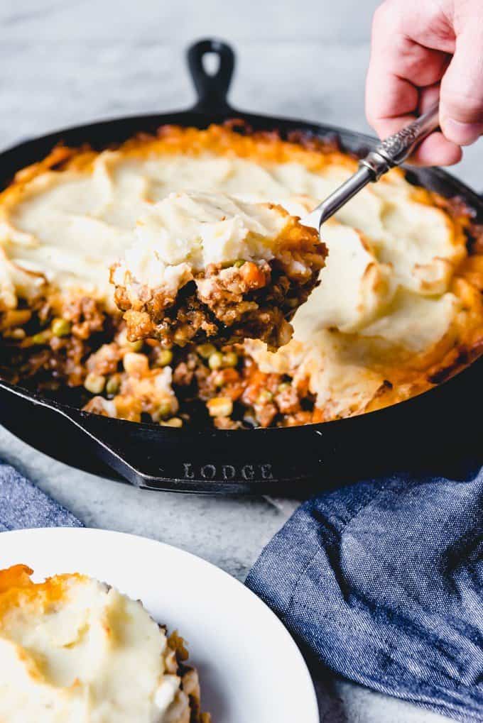 a hand holding a spoonful of shepherds pie above the cast iron skillet