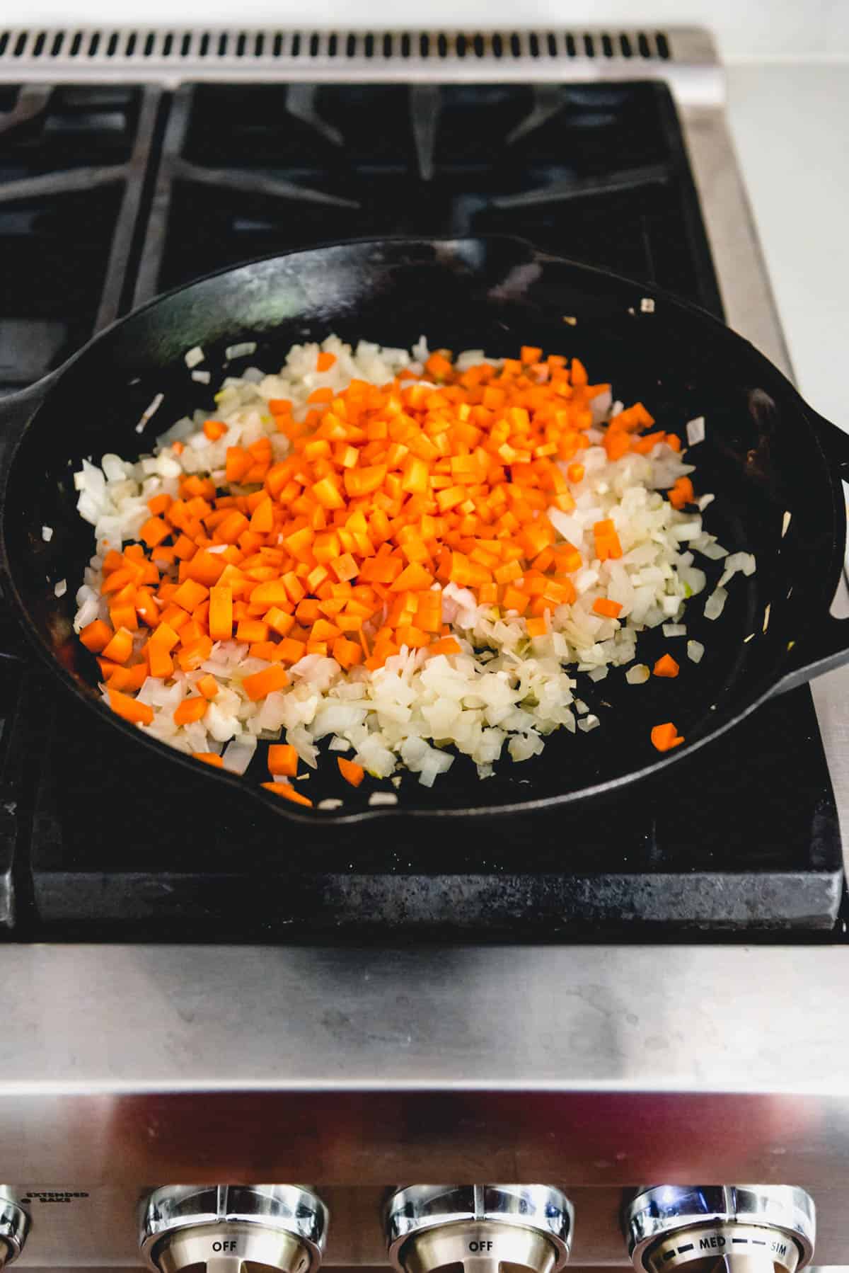 Adding diced carrots to a pan of sauteed onions in a cast iron skillet.