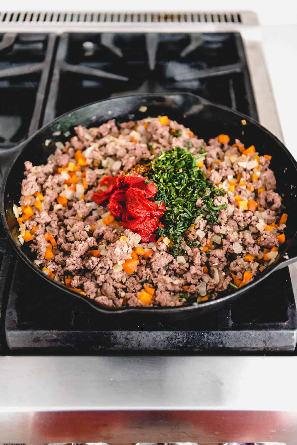 Adding tomato paste and other ingredients to browned meat in a cast iron skillet.