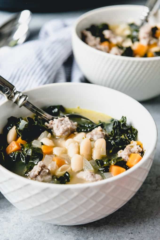An image of two white bowls of healthy bean soup with kale, Italian sausage, carrots, and onions, in a savory pesto infused chicken broth.