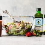 a large glass bowl filled with Spring Salad with Strawberry Balsamic Vinaigrette in a shaker to the side and bottles of olive oil on the other side