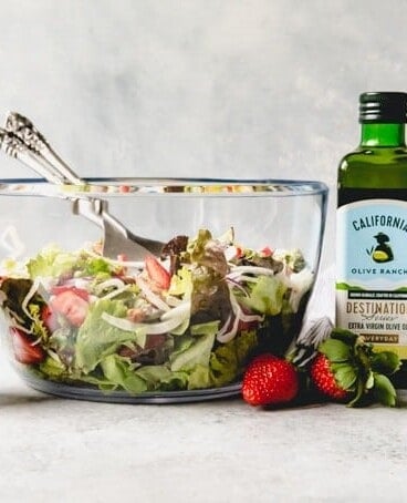 a large glass bowl filled with Spring Salad with Strawberry Balsamic Vinaigrette in a shaker to the side and bottles of olive oil on the other side