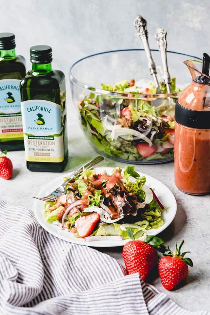 a white plate with salad topped with strawberry balsamic vinaigrette a jar of strawberry balsamic vinaigrette to the side next to a large bowl of salad and bottles of olive oil