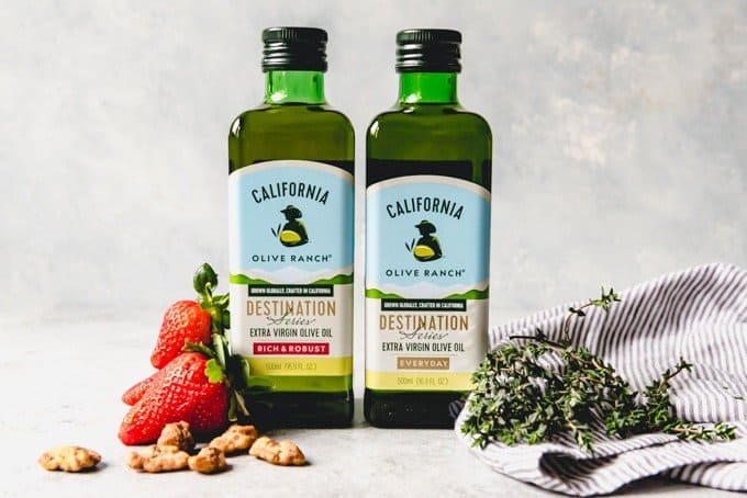 An image of two bottles of California Olive Ranch Extra Virgin Olive Oil with strawberries, candied walnuts, and fresh thyme next to them.