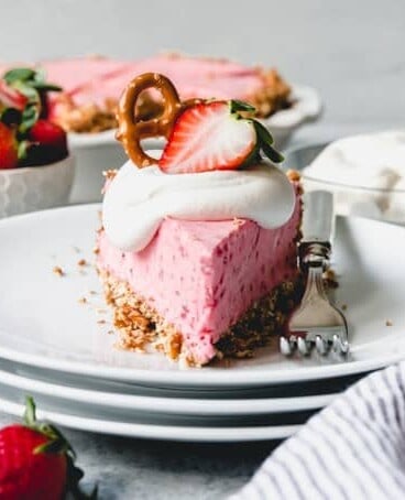 a stack of white plates with fresh strawberries to the side and a slice of strawberry pretzel icebox pie on top