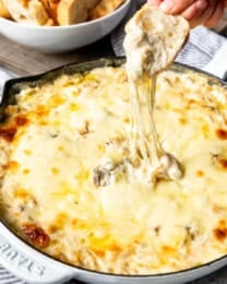 A hand pulling a slice of bread out of the philly cheese steak dip revealing the stringy cheesy goodness.