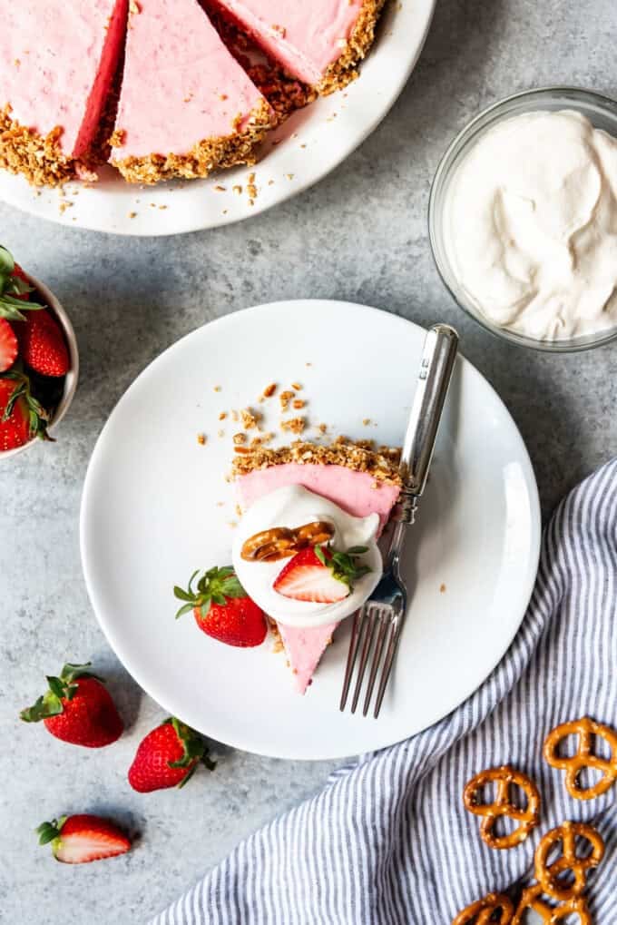 an aerial view of a slice of Strawberry Pretzel Icebox Pie on a white plate with a fork with fresh strawberries and pretzels to the side