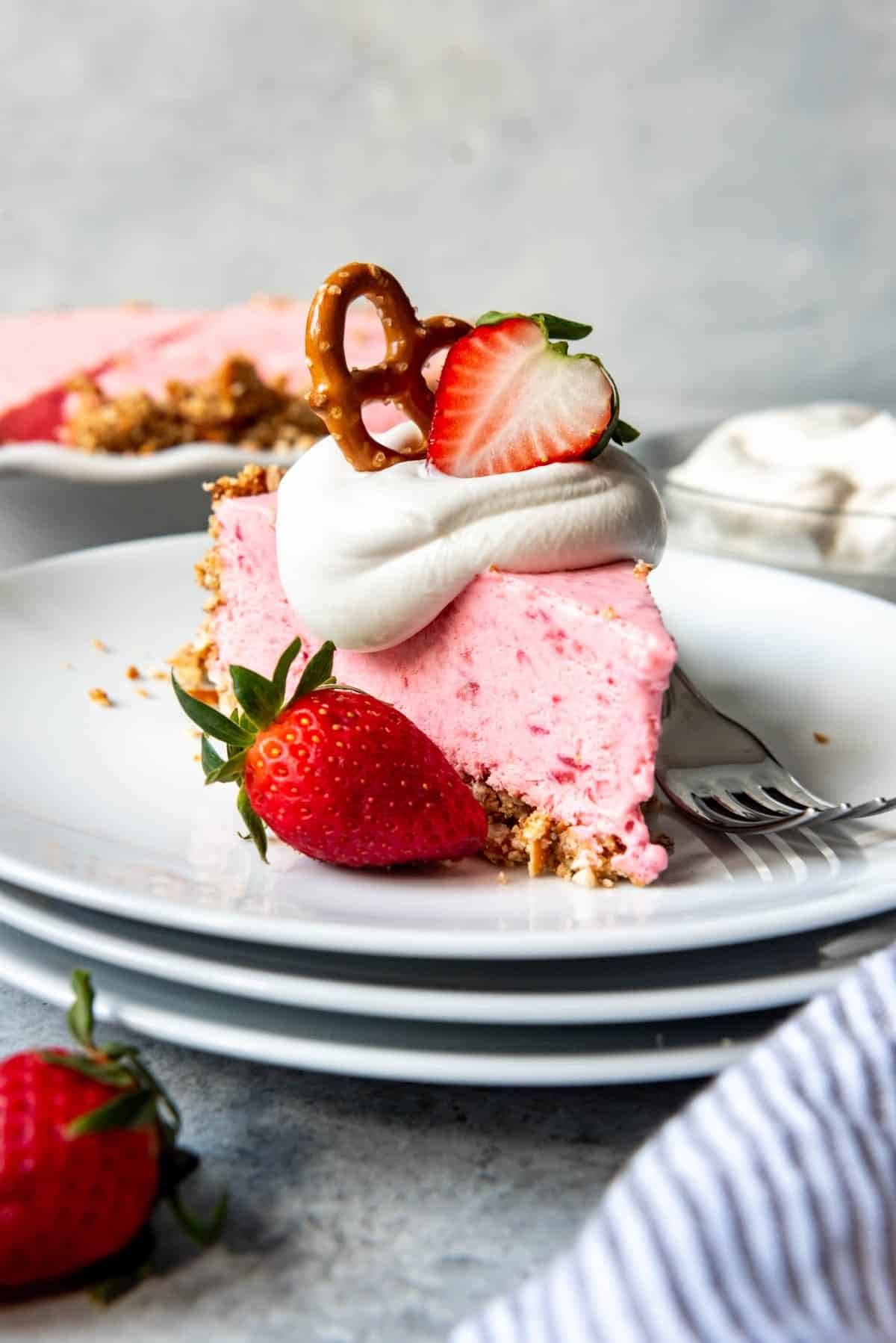 a pretzel and strawberries garnsihing a slice of Strawberry Pretzel Icebox Pie on a stack of white plates