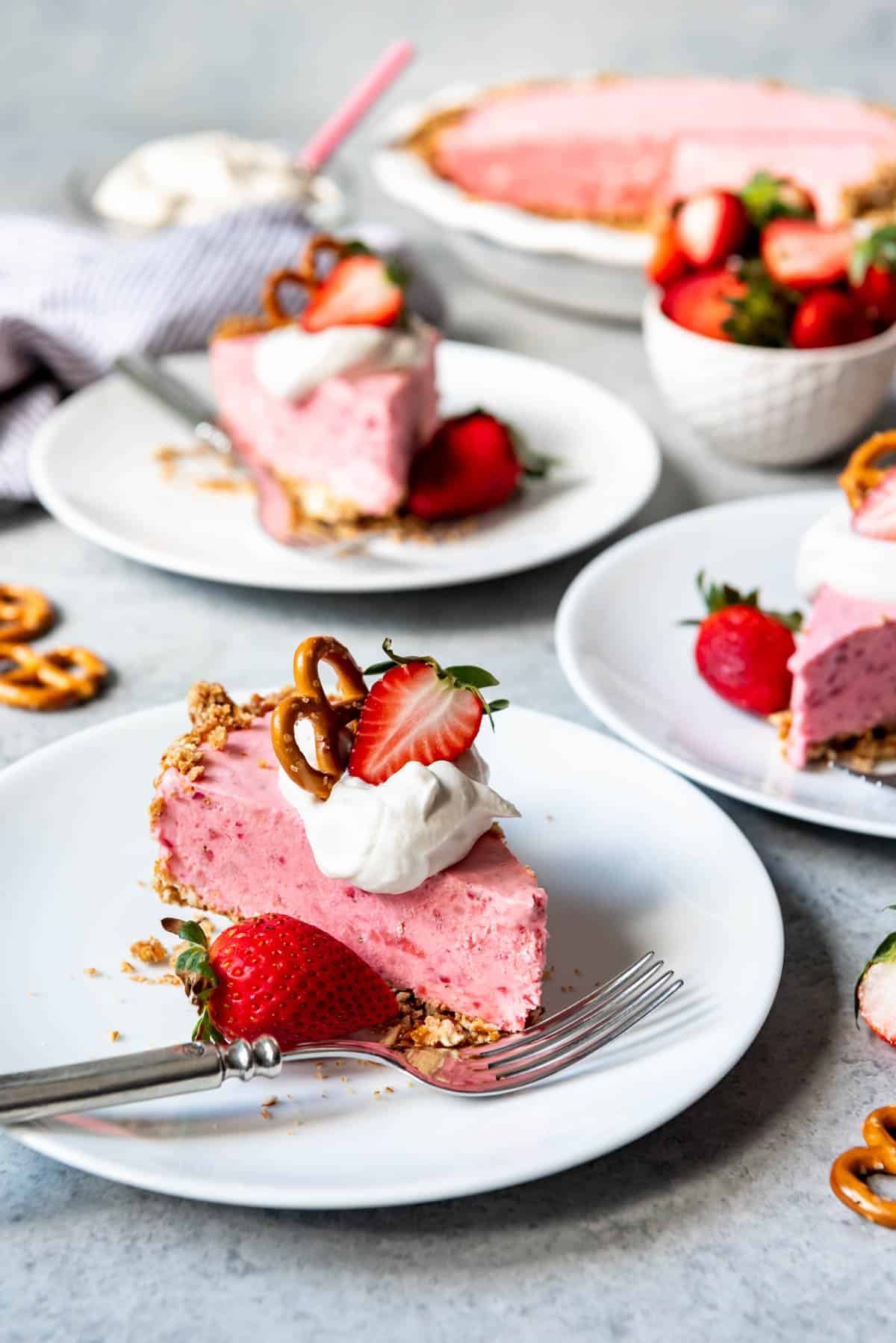 An image of a strawberry pie recipe with a pretzel crust, sliced and served on white plates with freshly whipped cream, pretzels, and strawberry halves on top.