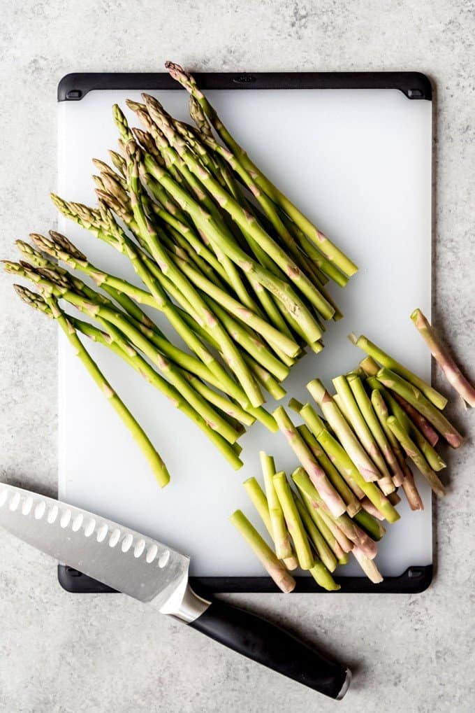 An image of a bunch of asparagus trimmed of it's tough ends on a cutting board with a chopping knife.