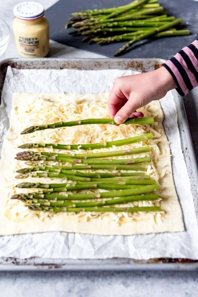 An image of a hand placing a stalk of asparagus on puff pastry sprinkle with cheese.