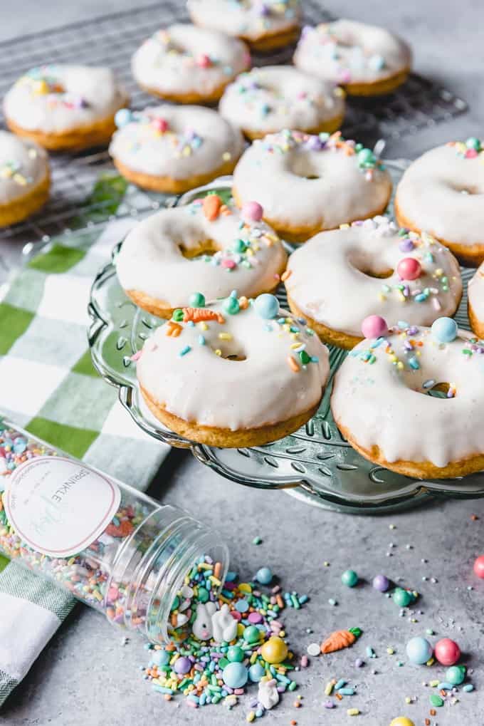 An image of baked carrot cake donuts on a green glass cake stand with Easter sprinkles on a cream cheese glaze.
