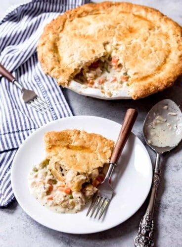 a white plate with a slice of chicken pot pie and a fork in front of a chicken pot pie missing a slice