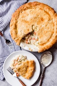 a white plate with a slice of chicken pot pie next to a used serving spoon and an almost full pie