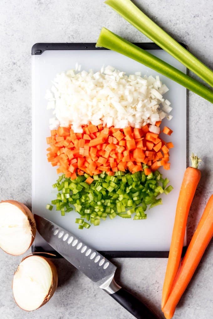 An image of diced carrots, celery, and onions on a cutting board with a knife.