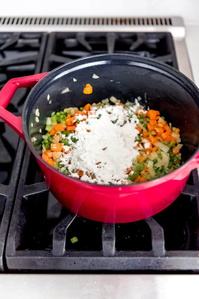 An image of flour sprinkled over softened vegetables in a large pan on the stove for thickening chicken pot pie filling.