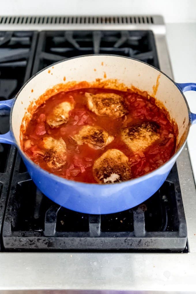 An image of seared chicken pieces stewing in a simple tomato sauce on the stovetop.