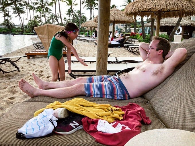a man with his arms behind his head on a chair at the beach and a daughter talking to him with her arm out
