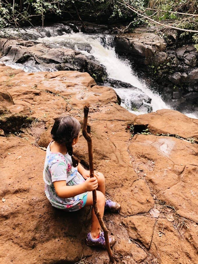 a girl sitting on a rock with her stick in hand looking at a small waterfall