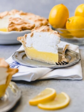 a slice of lemon meringue pie on a gray plate with a fork and sliced and whole lemons on the right with more pie on the left