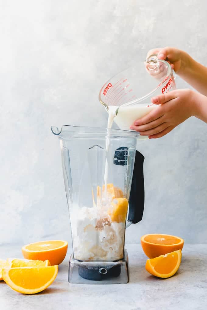 hands pouring liquid from a measuring glass into a blender with sliced oranges around it