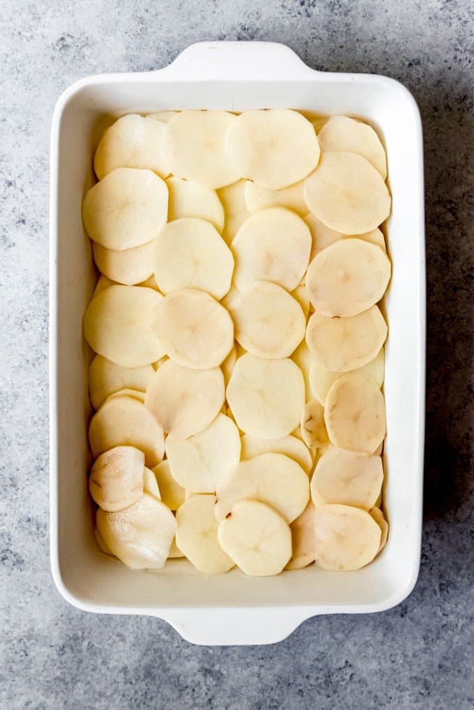 An image of thinly sliced potatoes arranged in layers on the bottom of a white casserole dish.