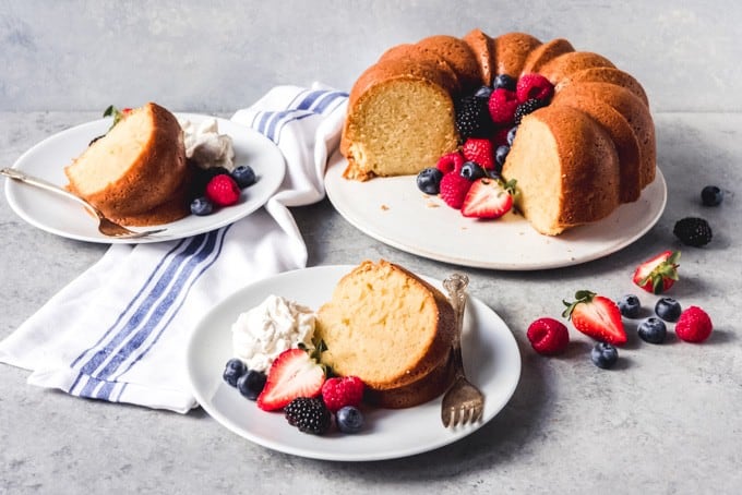 An image of slices of sour cream pound cake on dessert plates with berries and whipped cream next to the remaining unsliced cake.