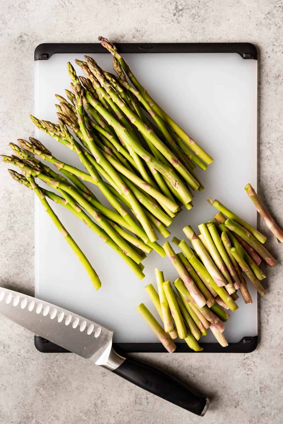 An image of a bunch of asparagus trimmed of it's tough ends on a cutting board with a chopping knife.