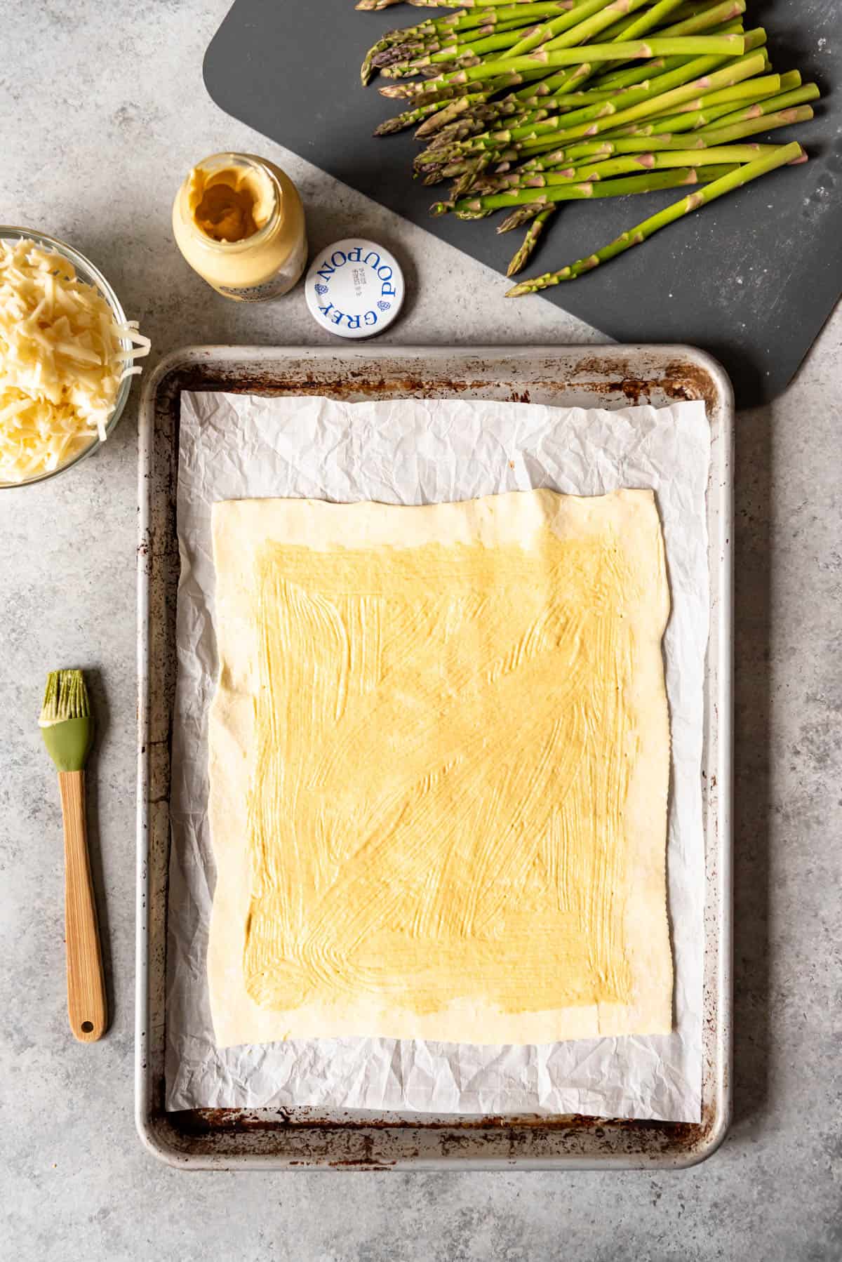 An image of puff pastry brushed with Dijon mustard on a baking sheet lined with parchment paper.