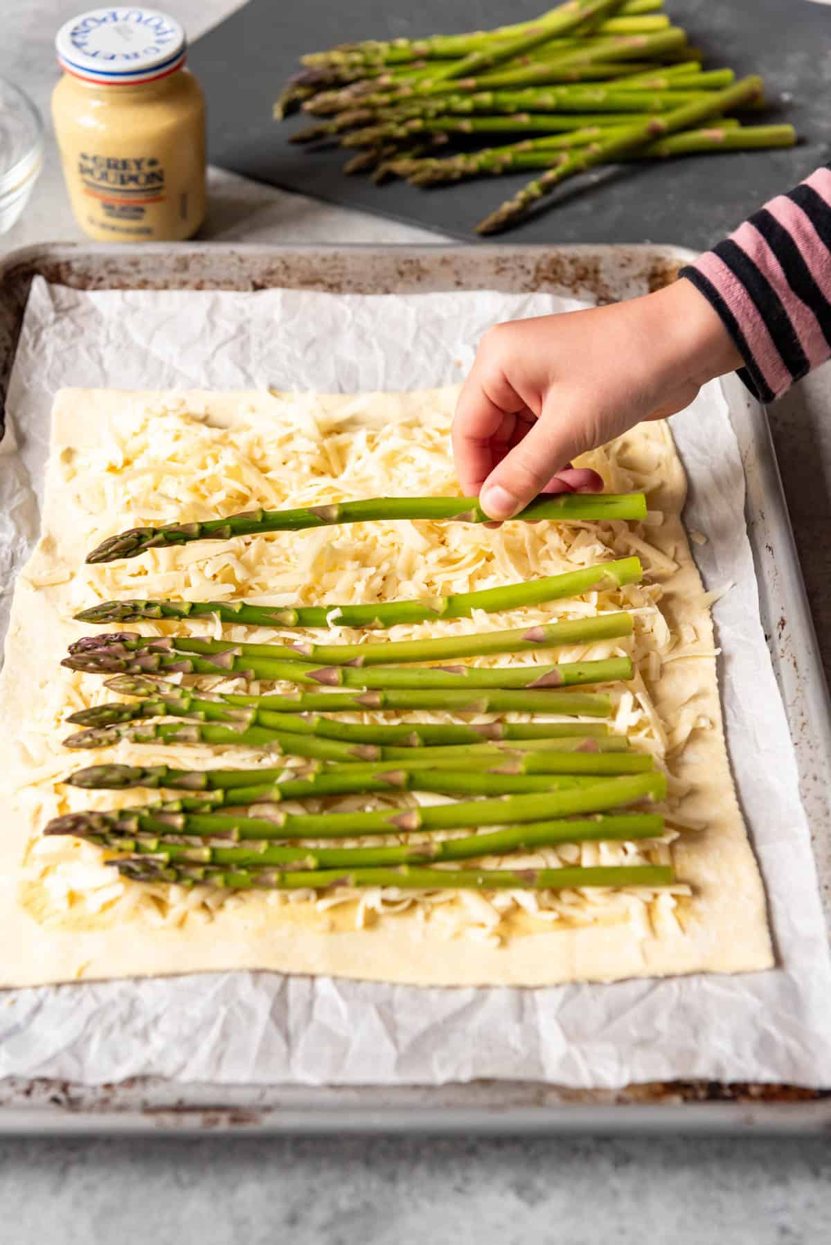 An image of a hand placing a stalk of asparagus on puff pastry sprinkle with cheese.