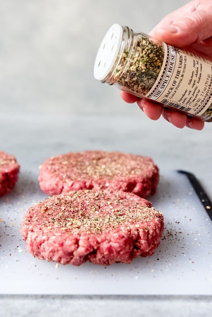 An image of a hand holding a shaker with hamburger seasoning blend in it to sprinkle over hamburger patties.
