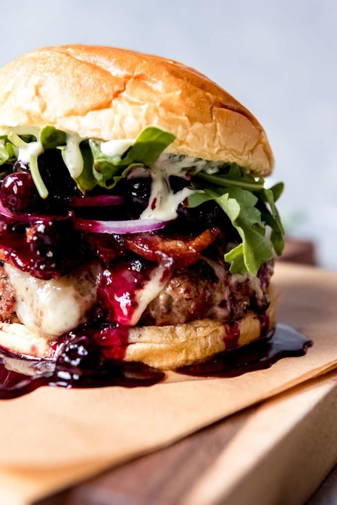 An image of a tall hamburger covered in cheese and blueberry balsamic sauce then topped with red onions, arugula, bacon, and basil aioli.