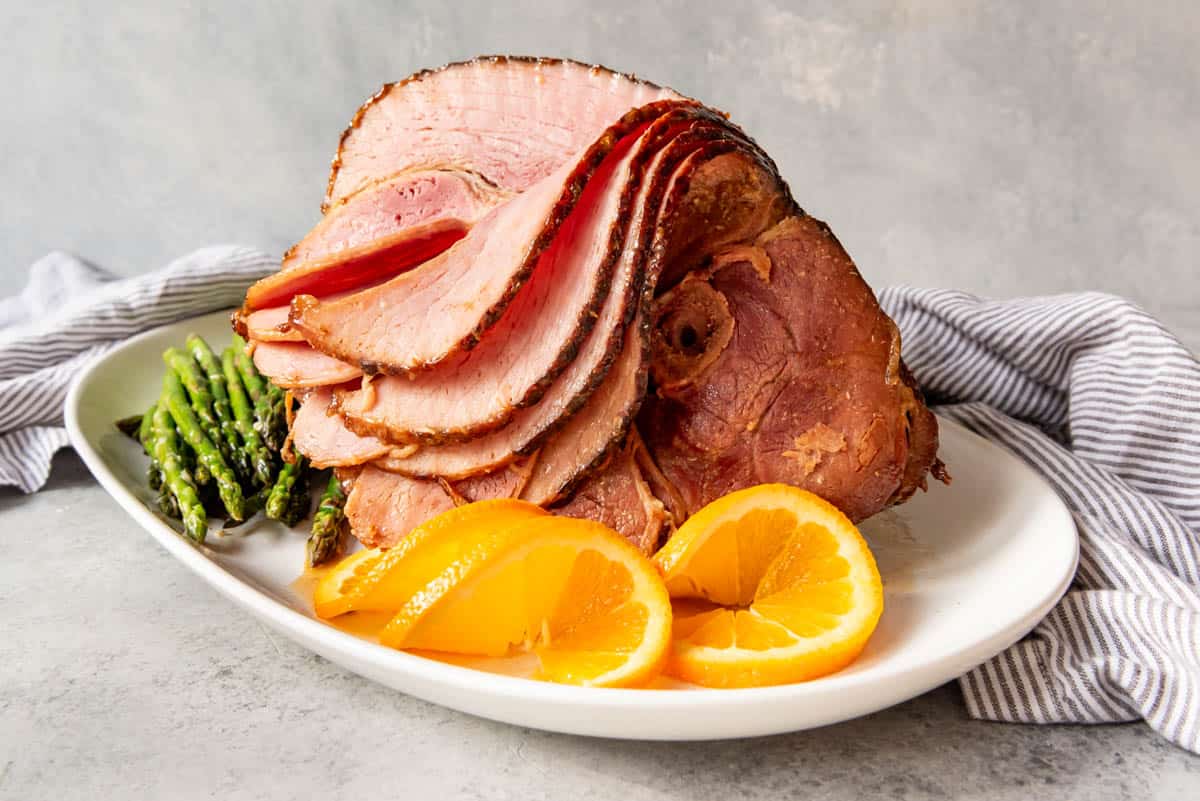 A spiral-cut glazed ham on a white serving platter with orange slices and asparagus.