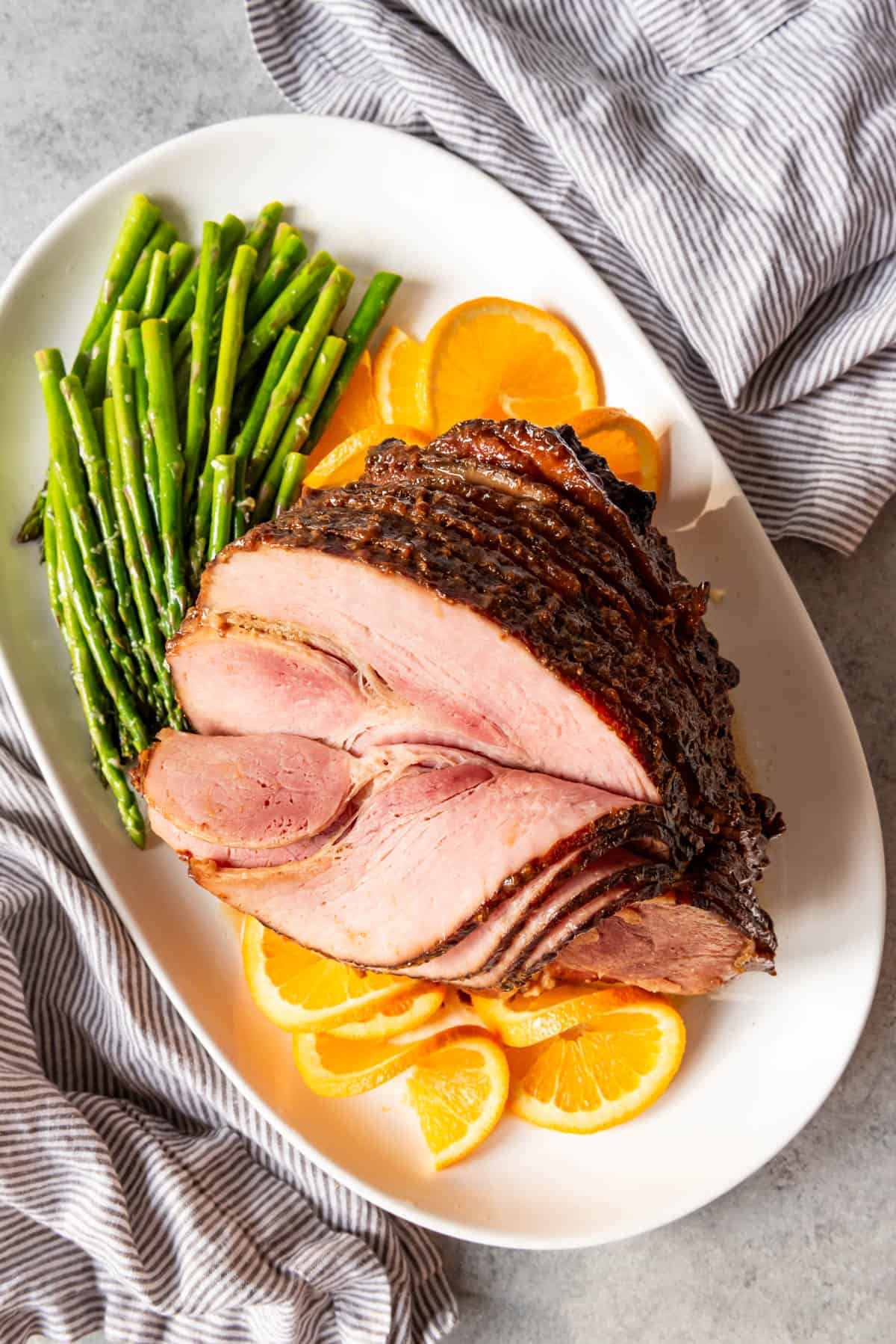A spiral cut ham with sliced oranges and asparagus on a white platter.
