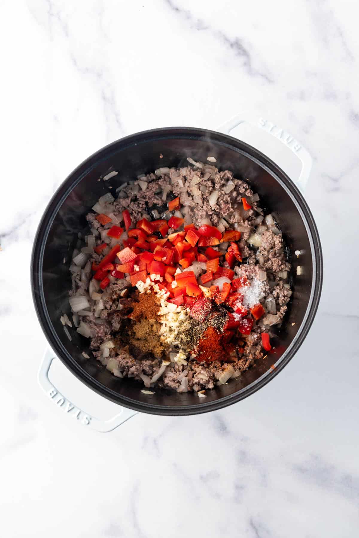 Adding diced veggies and spices to browned ground beef in a large dutch oven.