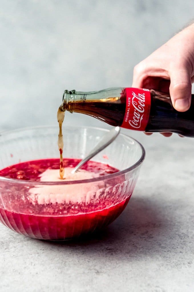 An image of a hand pouring a bottle of coca-cola into a bowl of cherry jello with fruit.