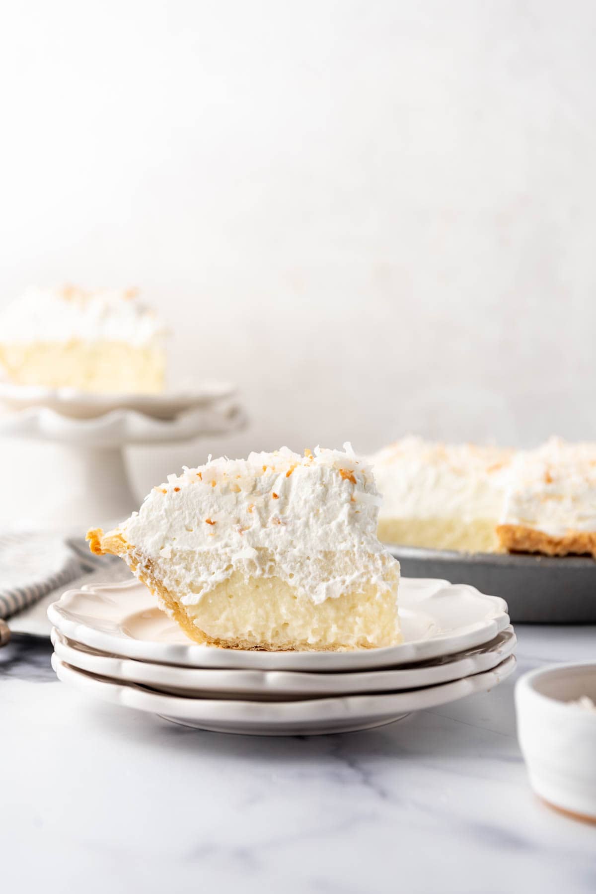 A slice of coconut cream pie on a stack of white dessert plates.