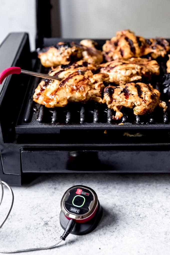 An image of chicken thighs on an indoor grill with the probe of a meat thermometer in one piece checking to see if it's done.