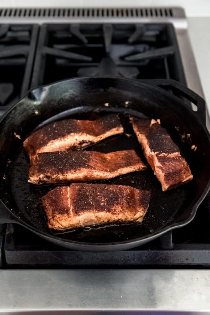 An image of blackened salmon fillets being cooked on the stove top in a cast iron skillet.